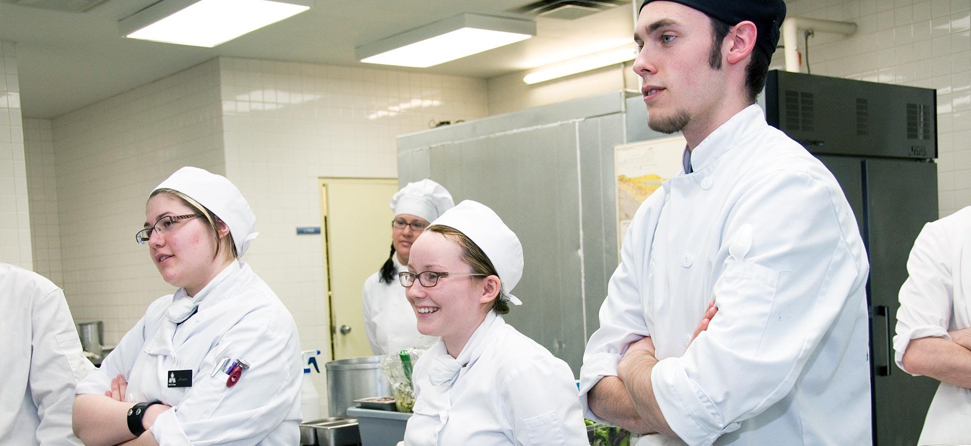 A group of culinary students listen to their instructor in one the ӣƵ culinary kitchens.