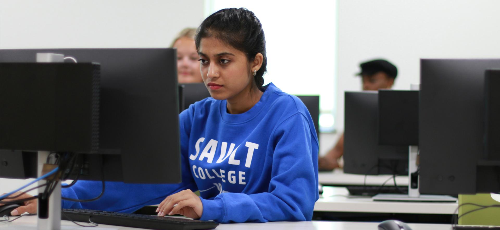 Student sitting at computer in class wearing blue ӣƵ sweater